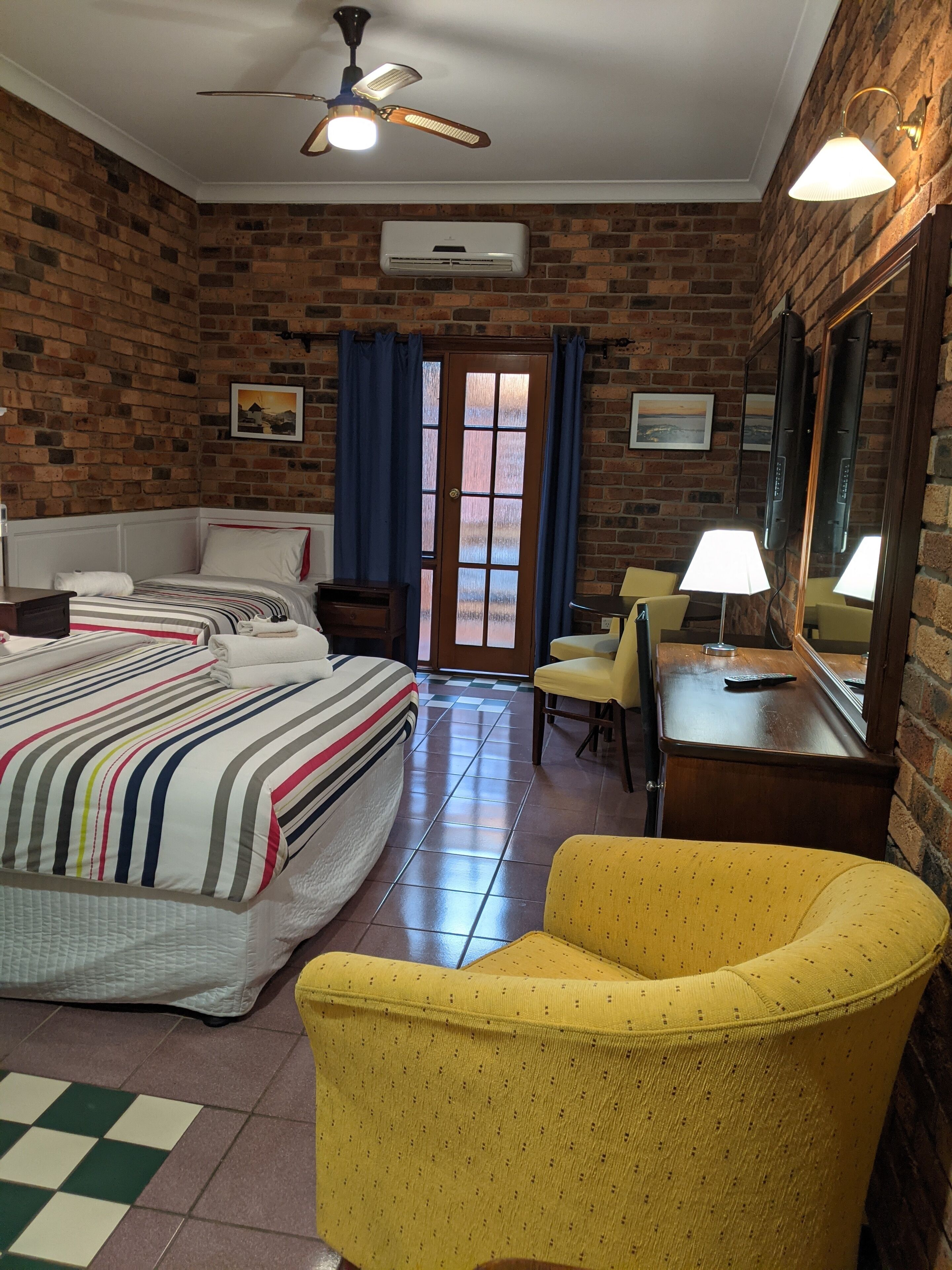 A hotel room with a bed, a yellow armchair, a desk with a lamp, a television, and a window with curtains.