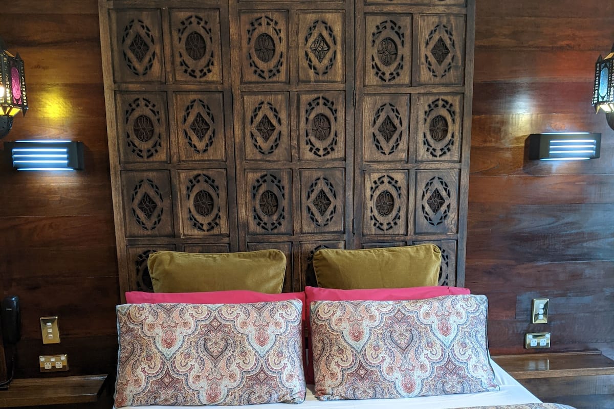 Un lit avec une couvre-lit à motifs et deux oreillers, une tête de lit en bois ornée de sculptures complexes, et deux appliques murales.