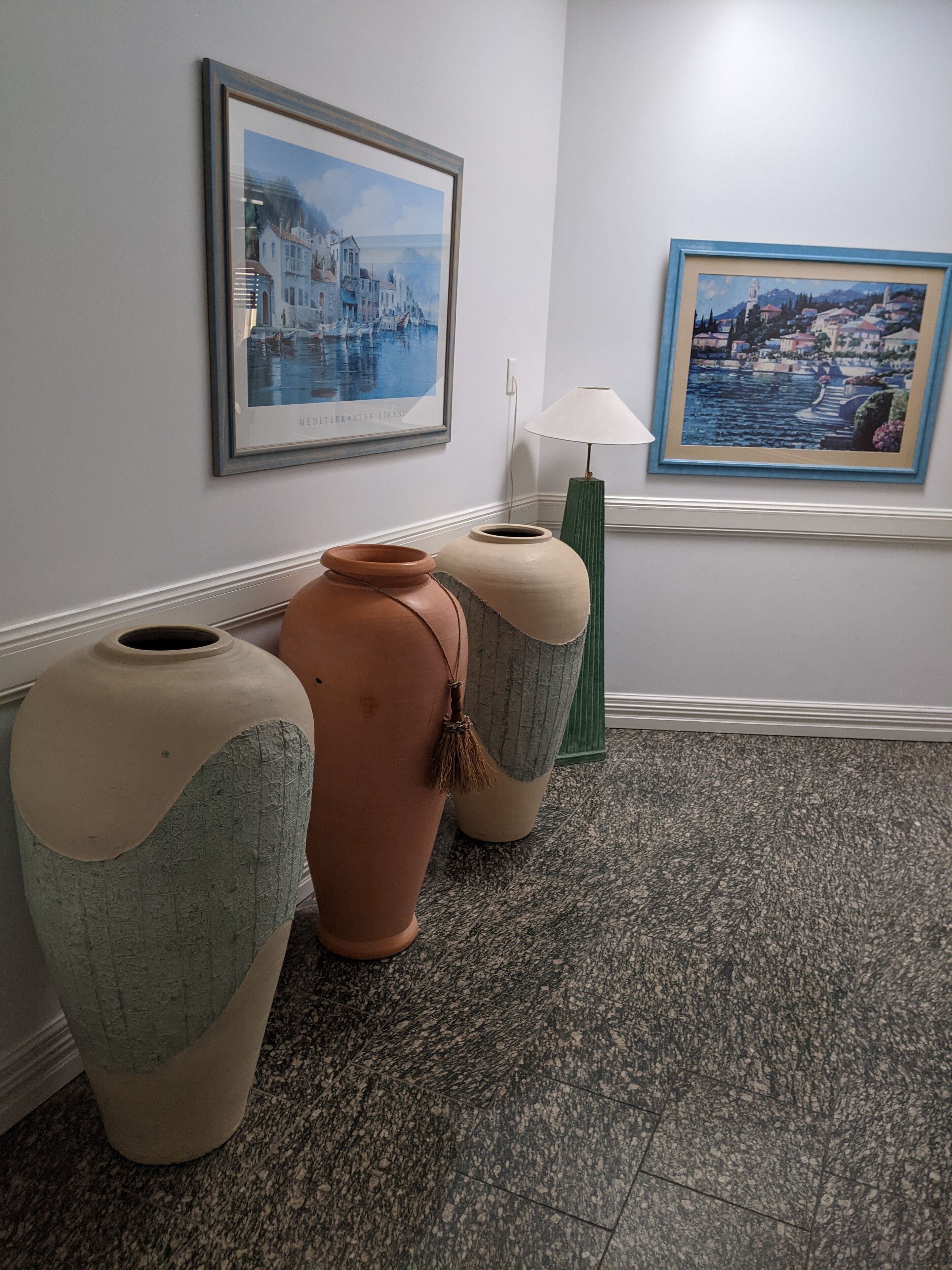 A hallway with two large vases, a framed painting of a Mediterranean town, and a lamp.