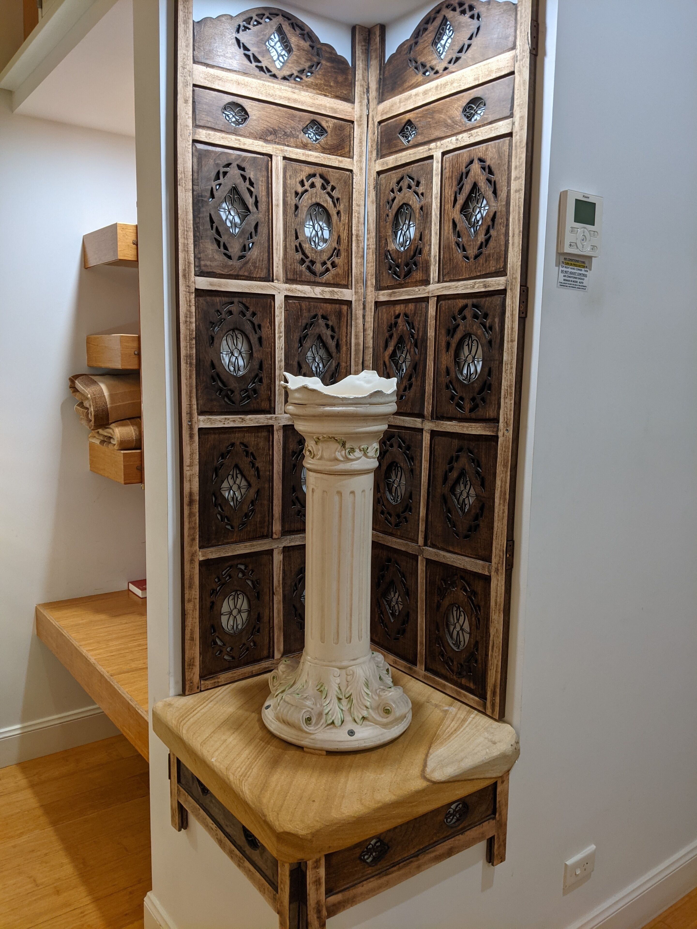A corner with a wooden cabinet featuring intricate carvings, a white pedestal, and a wooden shelf with books.