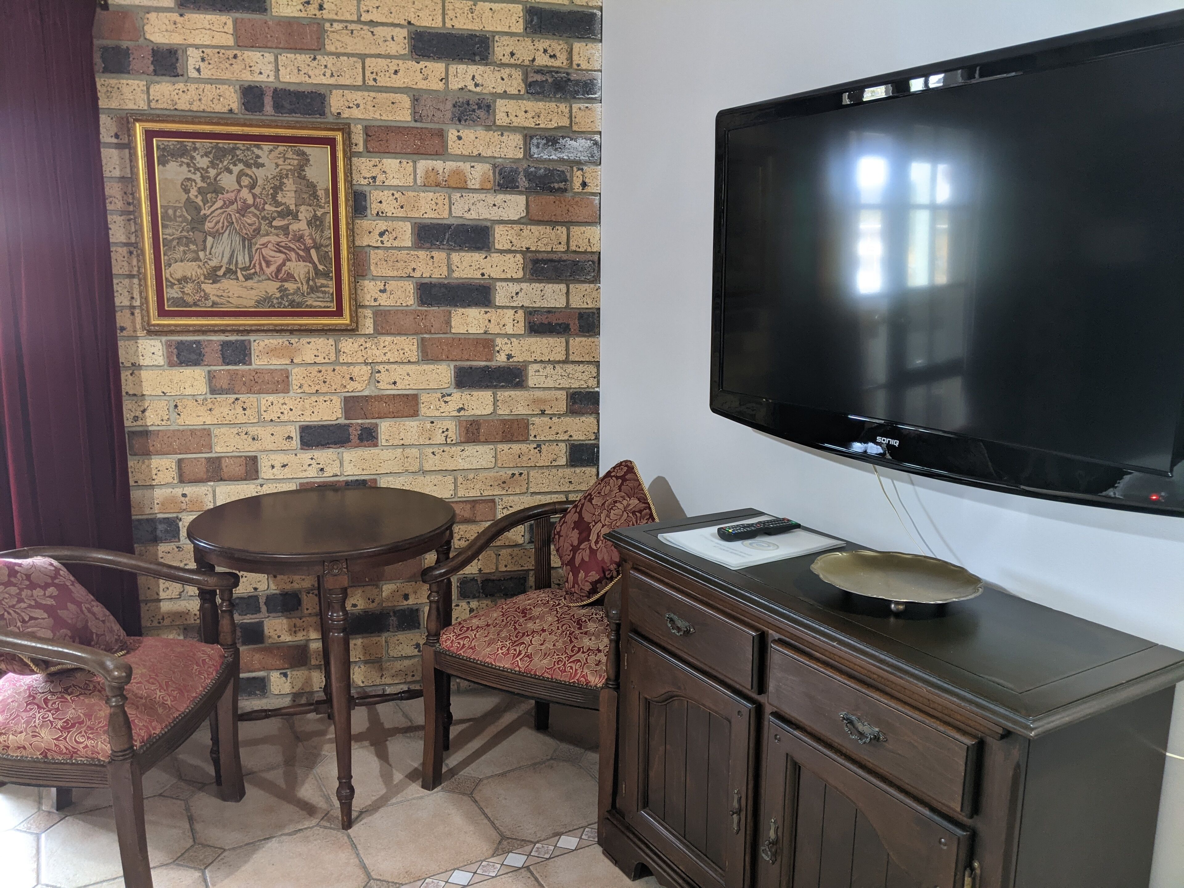 A room with a brick wall, a large flat-screen TV, a wooden cabinet, a table, and two chairs.