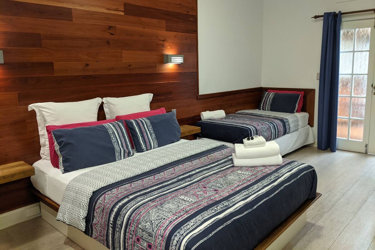 Une chambre d’hôtel avec deux lits, des lambris en bois aux murs, une fenêtre avec des rideaux et un panneau indiquant le mot « Soyez ».