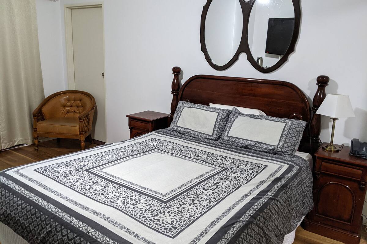 Une chambre avec un grand lit, une tête de lit en bois, une table de chevet avec une lampe, une chaise et un miroir au mur.