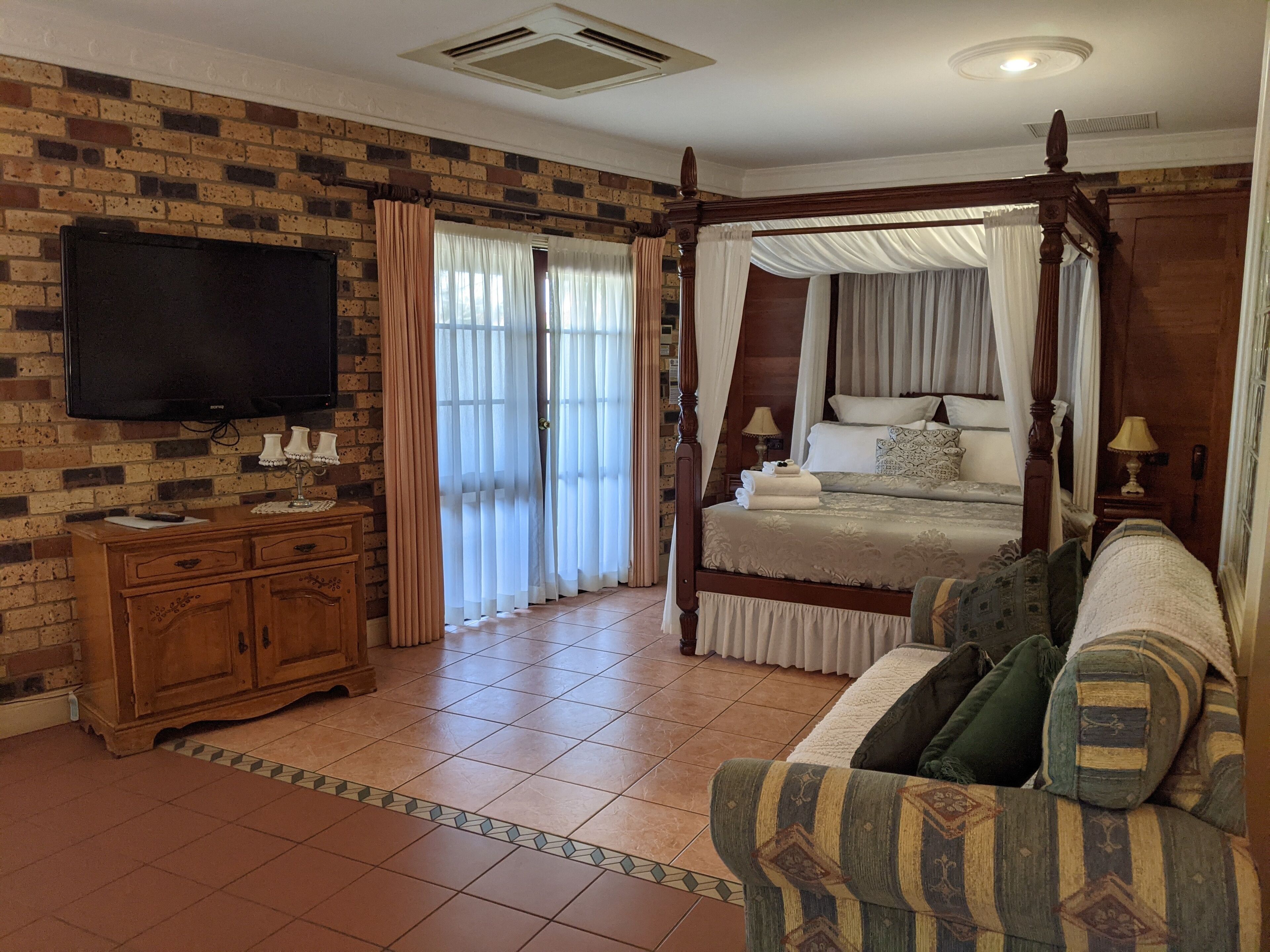 Un dormitorio con cama con dosel, televisión montada en la pared de ladrillo, cómoda de madera y sofá estampado.
