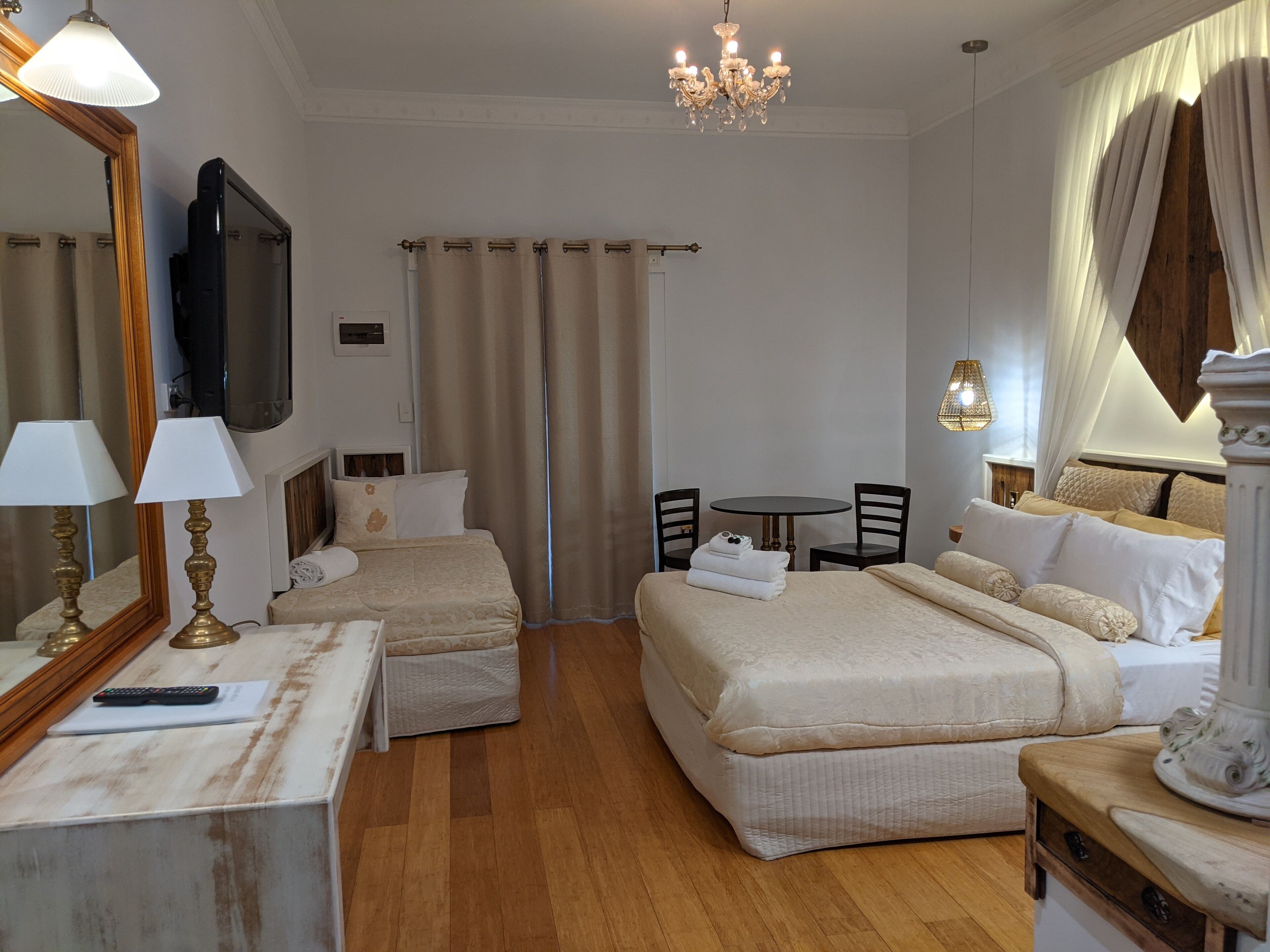 A hotel room with two beds, a TV, a chandelier, a dining table with chairs, and a large window with curtains.