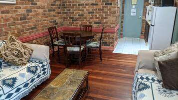 A cozy room with a brick wall, a dining table with chairs, a sofa, and a small kitchen area with a refrigerator.