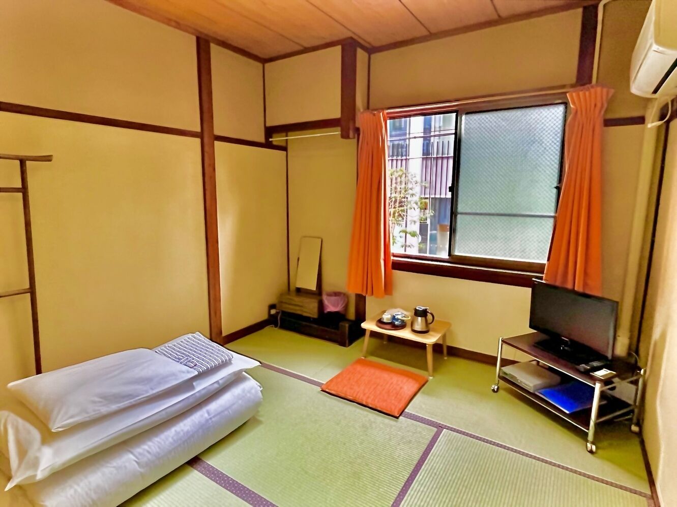 A room with tatami flooring, a bed, a TV on a stand, a small table with a kettle, and a window with curtains.