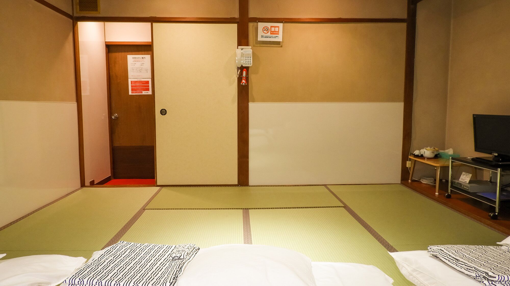 A room with tatami mats, a bed with a patterned blanket, a television on a stand, and a door with a fire alarm.