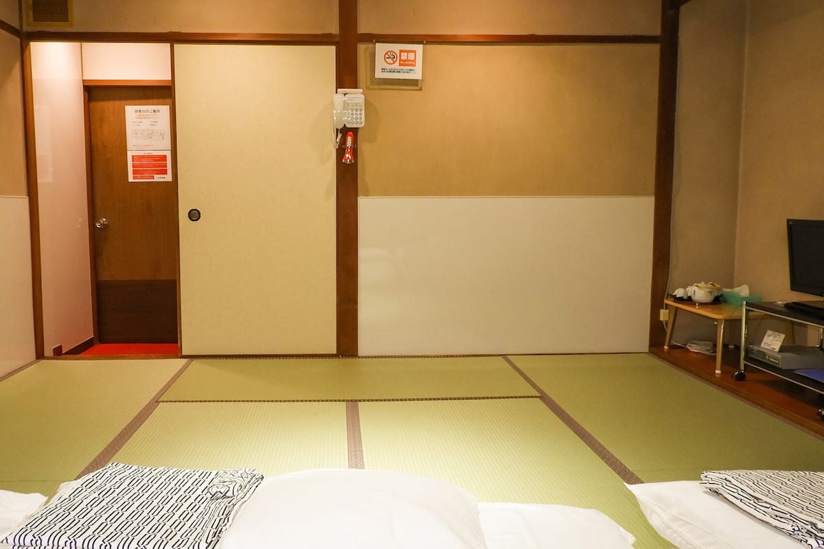 A room with tatami mats, a bed with a patterned blanket, a television on a stand, and a door with a fire alarm.