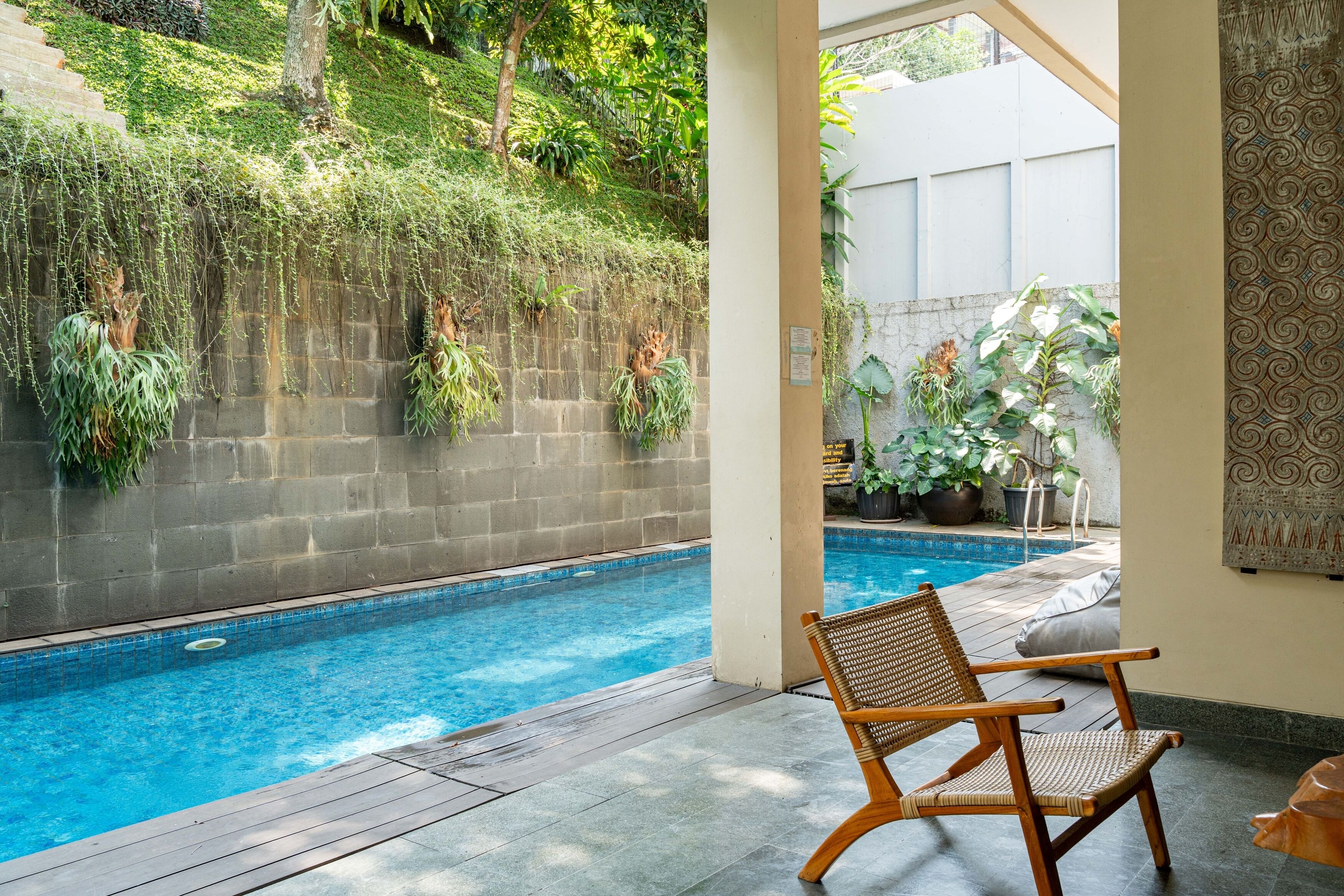 A pool area with a wooden chair, a stone wall with hanging plants, and a view of a building in the background.