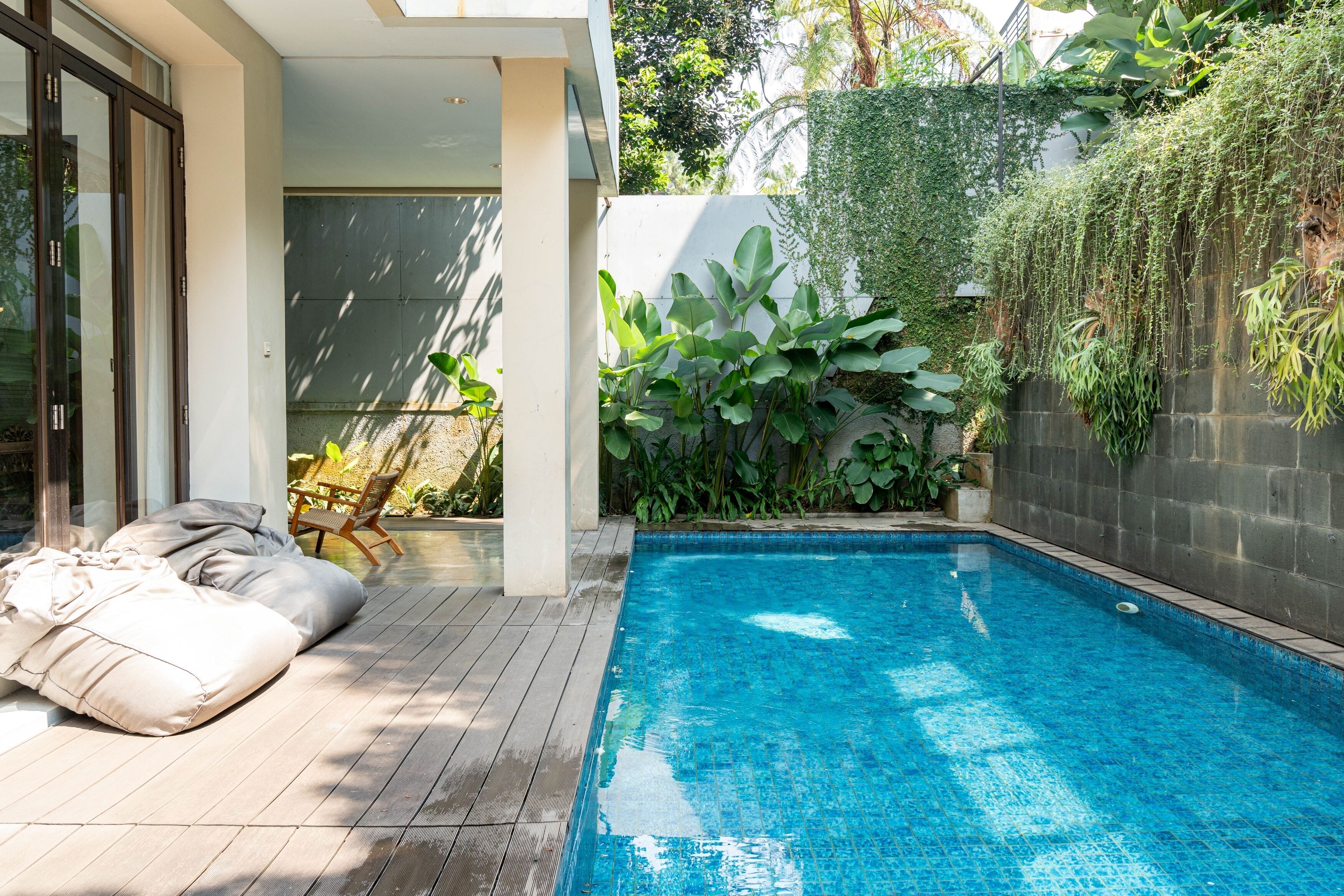 A modern outdoor pool area with a wooden deck, a seating area with cushions, and lush greenery.