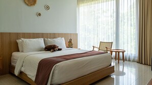 Ein Hotelzimmer mit einem großen Bett, einem Nachttisch, einem Sessel und Blick ins Freie durch transparente Vorhänge.