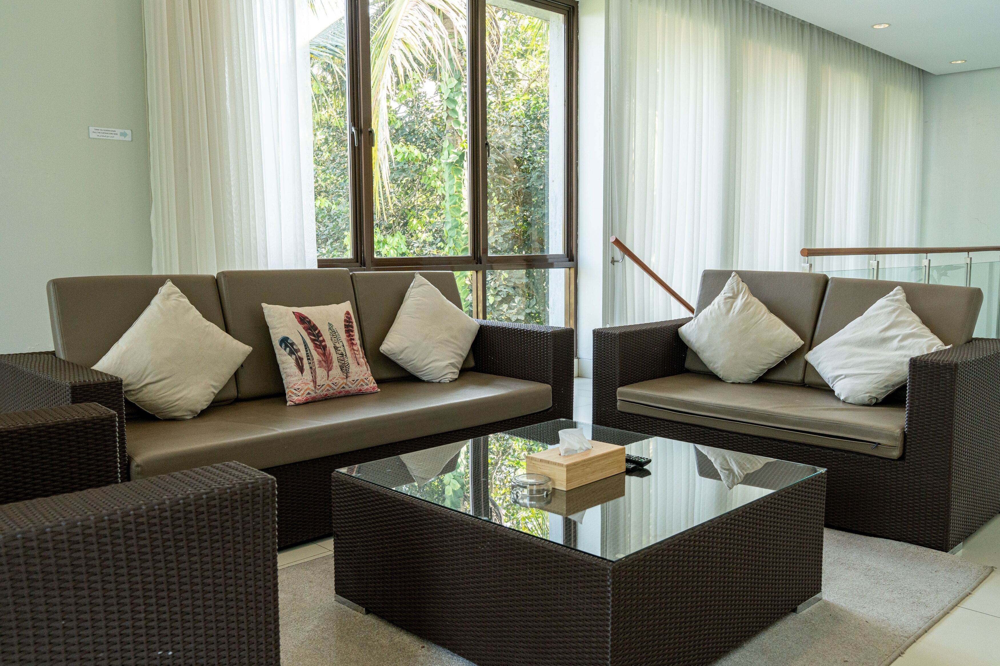 A modern living room with a sofa set, a glass coffee table, and large windows with curtains.