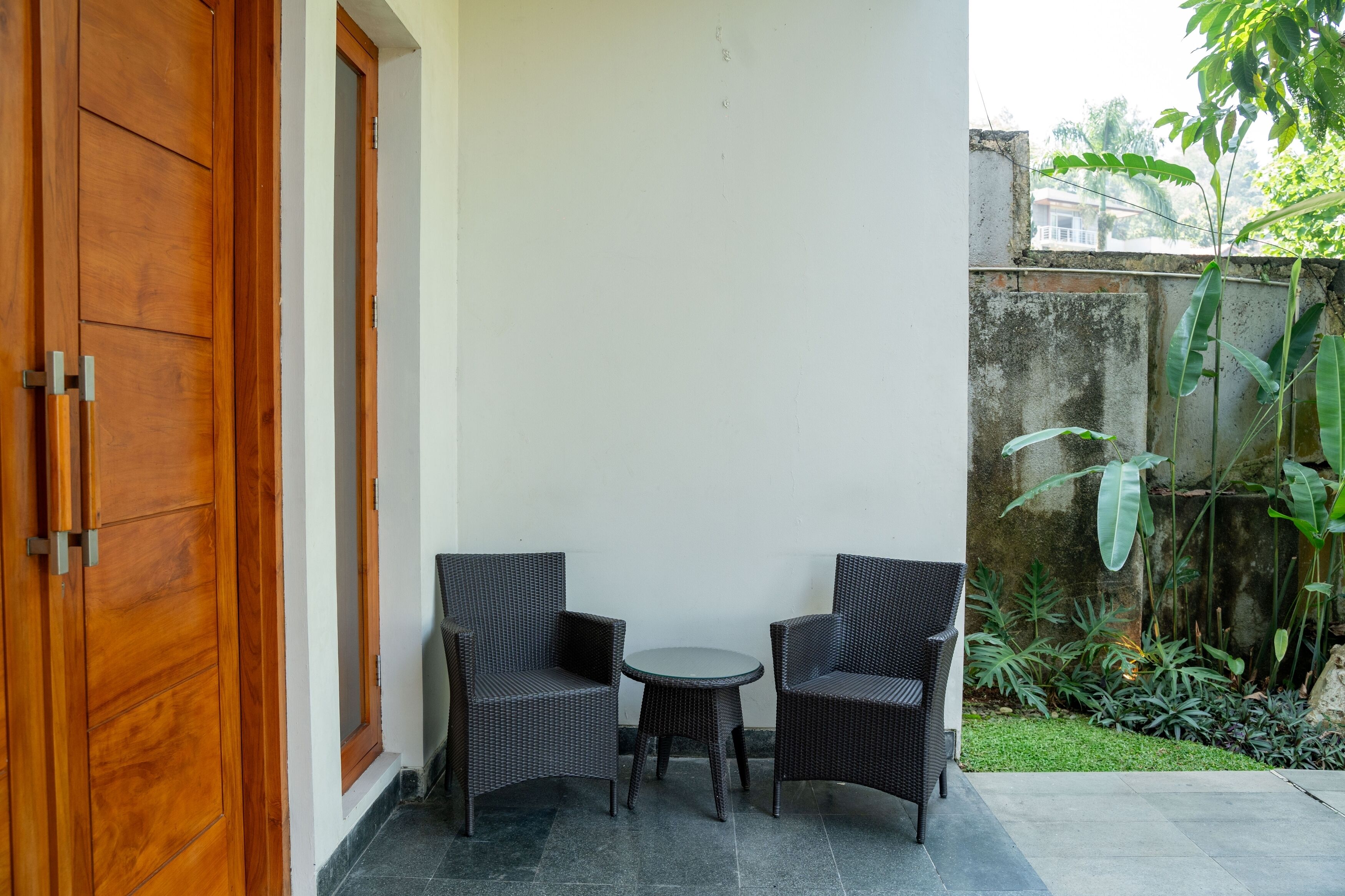 A small patio with two wicker chairs and a round table, a wooden door, and a concrete wall with greenery.