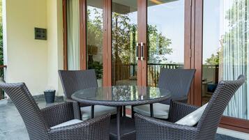 Una mesa redonda de vidrio con sillas de mimbre, una puerta corrediza de cristal y vista a la vegetación exterior.