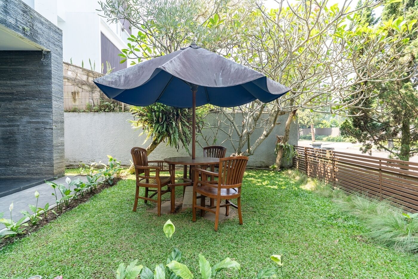 A patio with a table and chairs under a blue umbrella, surrounded by greenery and a fence.