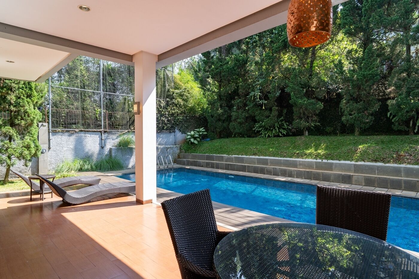 A modern outdoor pool area with a clear view of the surrounding greenery, a covered seating area, and a hanging light fixture.