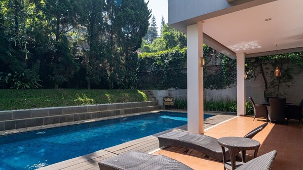 Ein Pool mit zwei Korbsesseln, einer überdachten Terrasse und Blick auf einen üppigen Garten.
