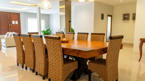 Un comedor con una gran mesa de madera rodeada de sillas de mimbre, un candelabro y vistas a una sala de estar con un sofá y cortinas.