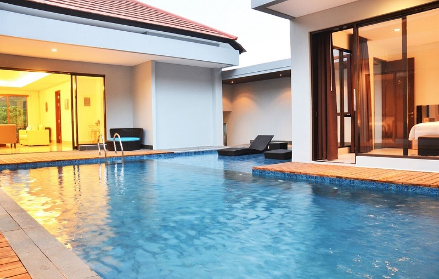 A swimming pool with a wooden deck, a glass door leading to a room, and a chair by the pool.