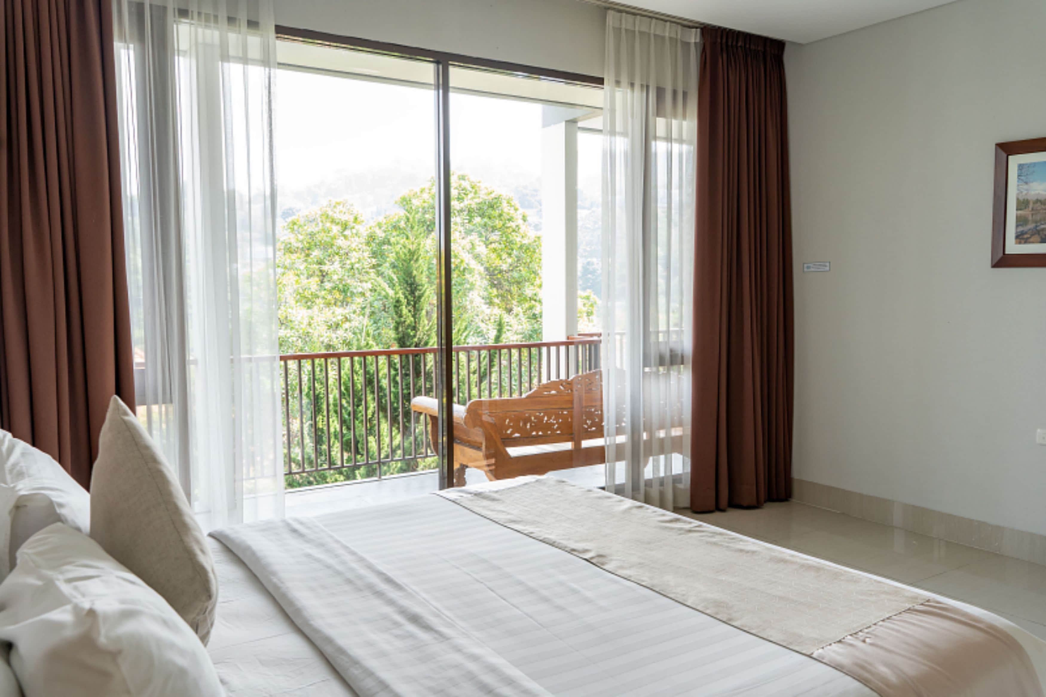 Ein Hotelzimmer mit einem großen Bett, einem Balkon mit einem Holzstuhl und Blick auf Grünfläche.
