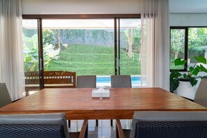 Un comedor con mesa y sillas de madera, una banca y vistas a un jardín exuberante y piscina.