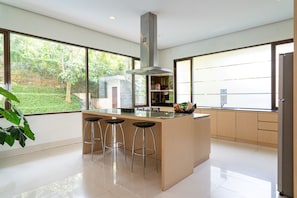 A modern kitchen with a central island, bar stools, and large windows offering a view of greenery. - Oyama City View 4BR Villa with a Heated Pool (Bandung)