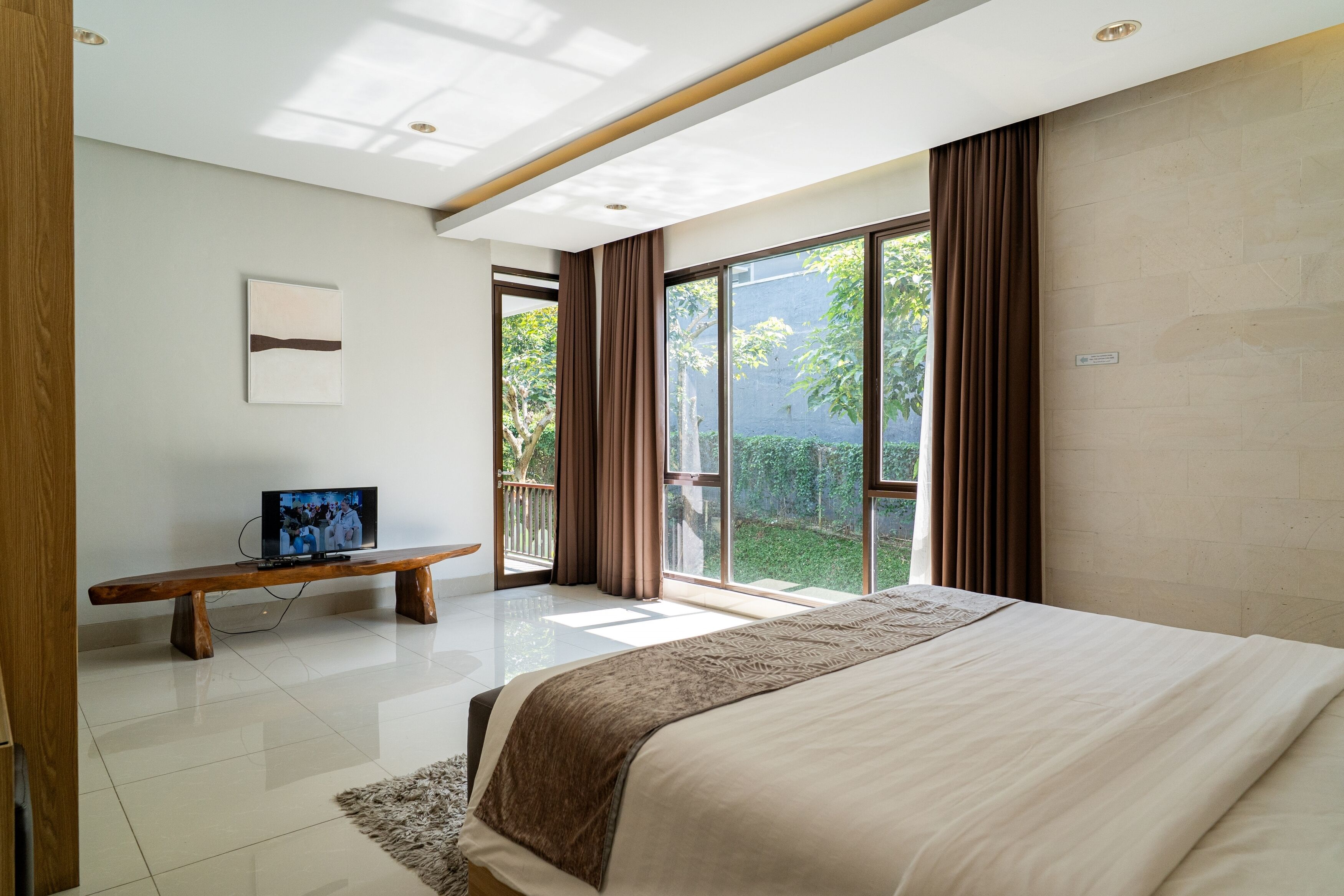 A modern bedroom with a large bed, a bench, a television, and a balcony with a view of greenery.