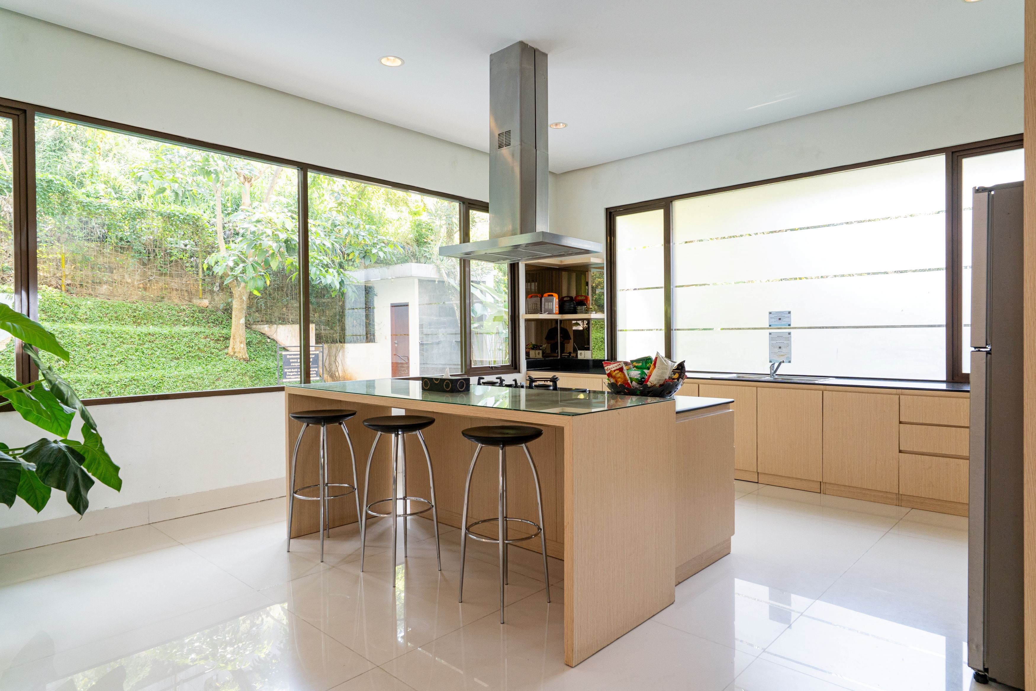 A modern kitchen with a central island, bar stools, and large windows offering a view of greenery.