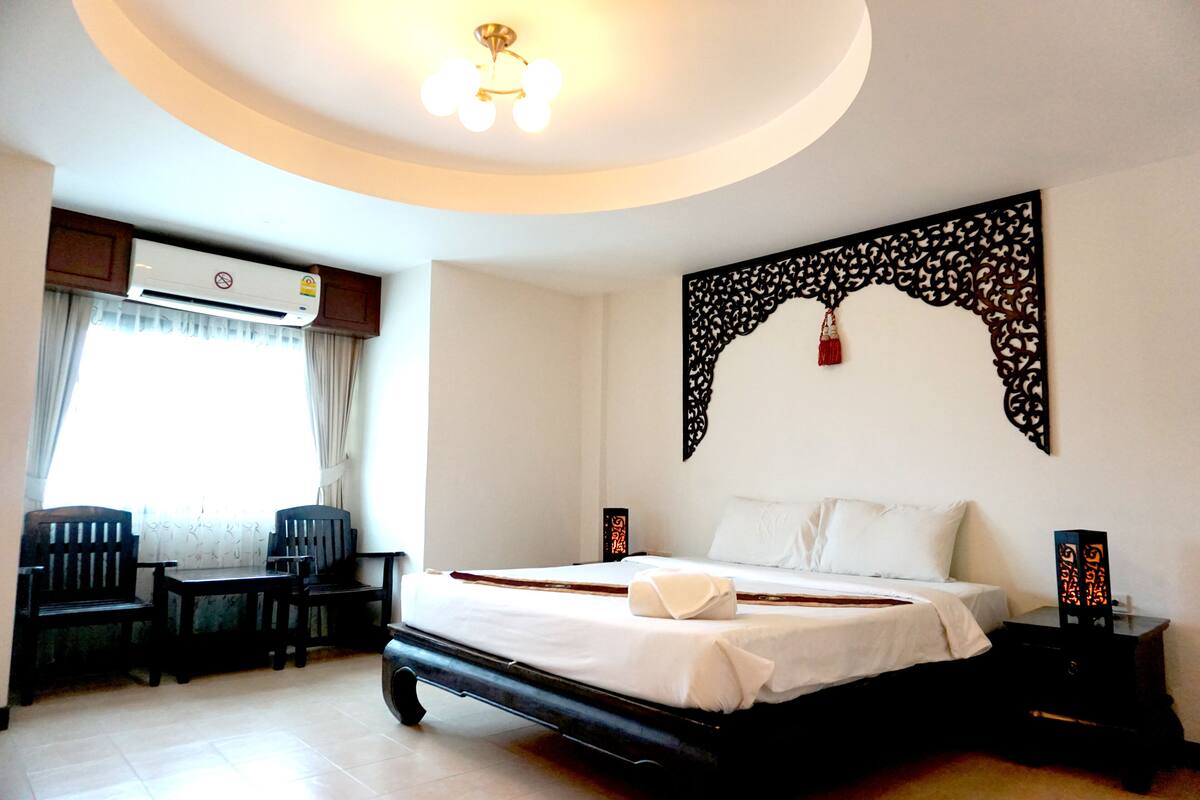 Hotelkamer met een bed, bureau, twee stoelen, airconditioning, een decoratief wandobject en een nachtkastje met lamp.