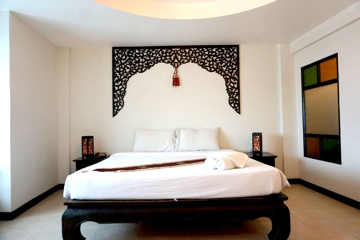 Een net opgemaakt bed met witte lakens, een decoratieve wandhanger erboven en twee nachtkastjes met lampen.