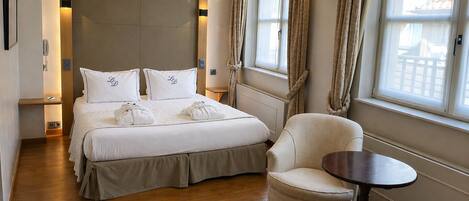 Habitación de hotel con una cama grande, una silla, una mesita y vistas al exterior por la ventana.