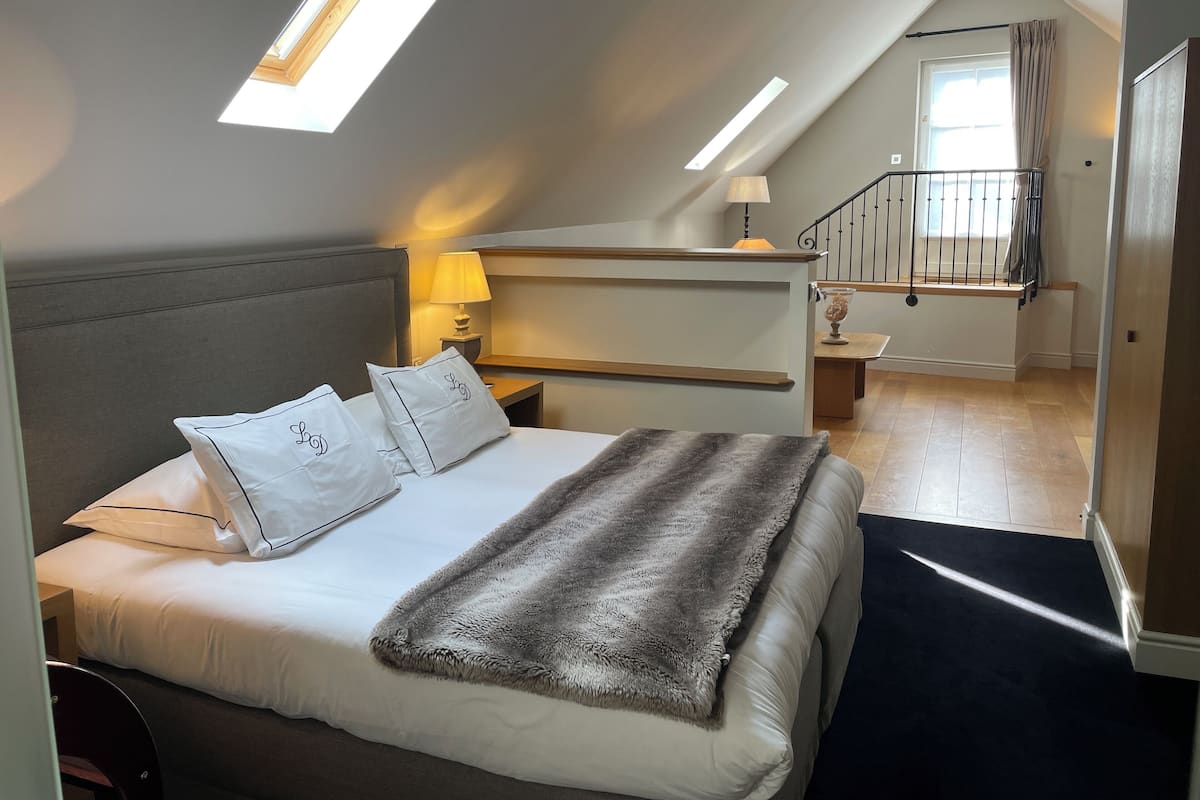 A bedroom with a bed, pillows, a bedside lamp, a window, a staircase, and a door.