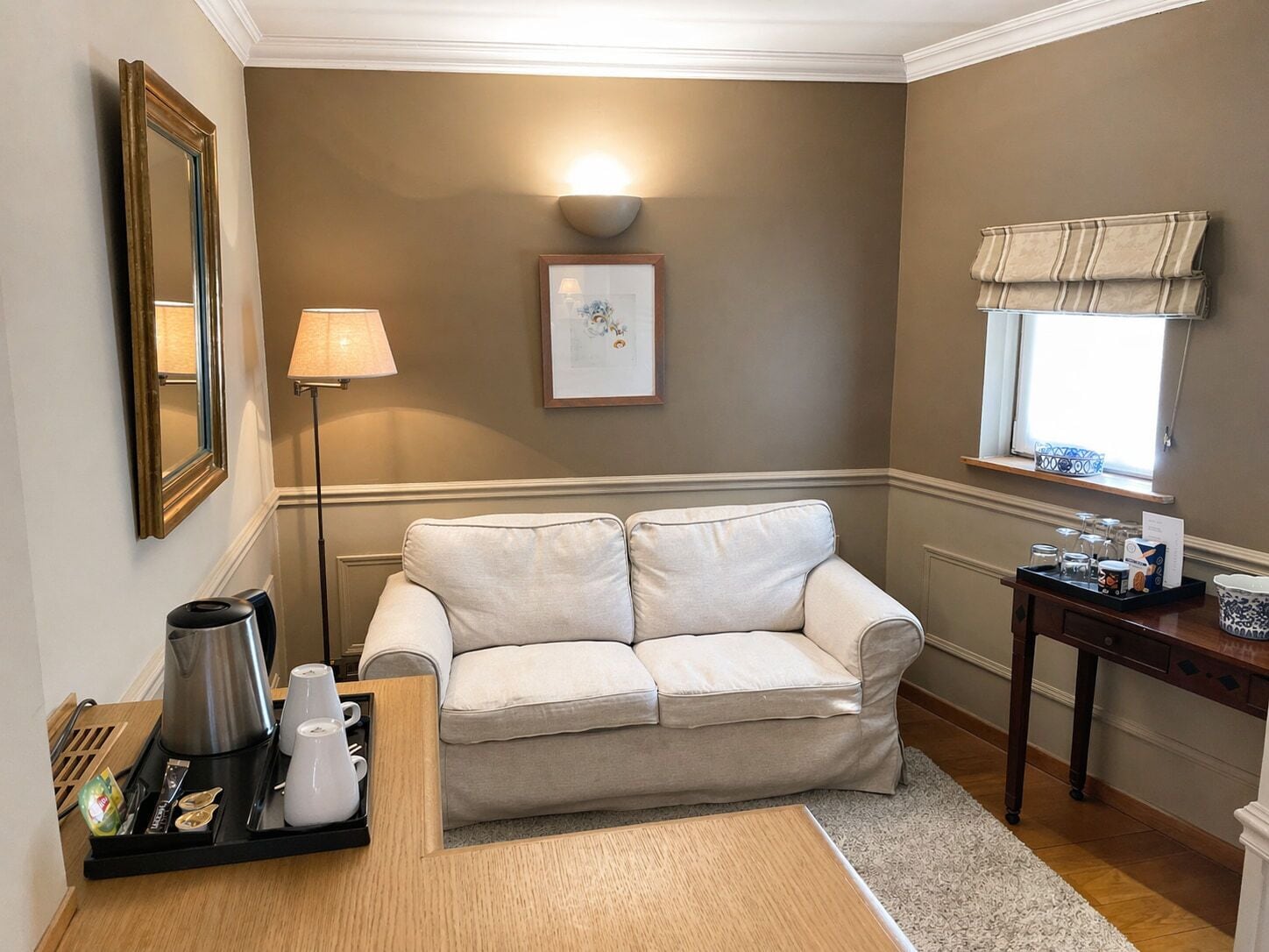 Una habitación con un sofá color crema, una estación de café, un cuadro enmarcado y una ventana con cortinas.