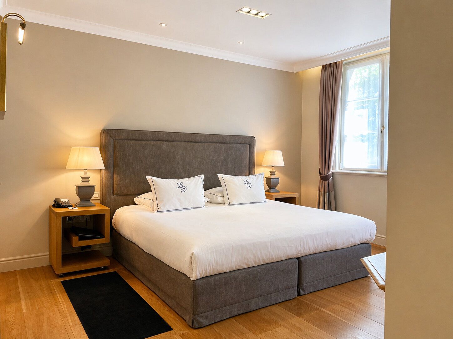 A bedroom with a large bed, two bedside tables with lamps, a window with curtains, and a wooden floor.