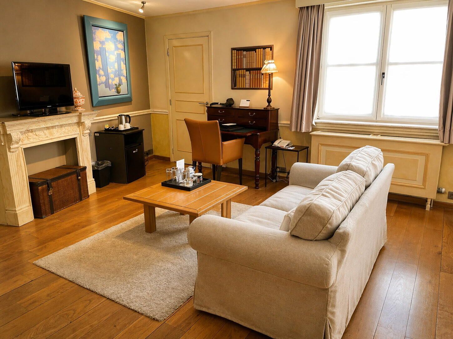Ein Wohnzimmer mit einer Couch, einem Couchtisch, einem Kamin, einem Fernseher, einem Schreibtisch, einem Bücherregal und einem Fenster.