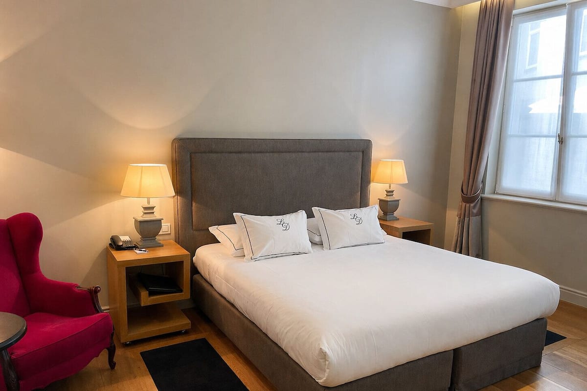 A hotel room with a large bed, two bedside tables with lamps, a red armchair, and a window with curtains.