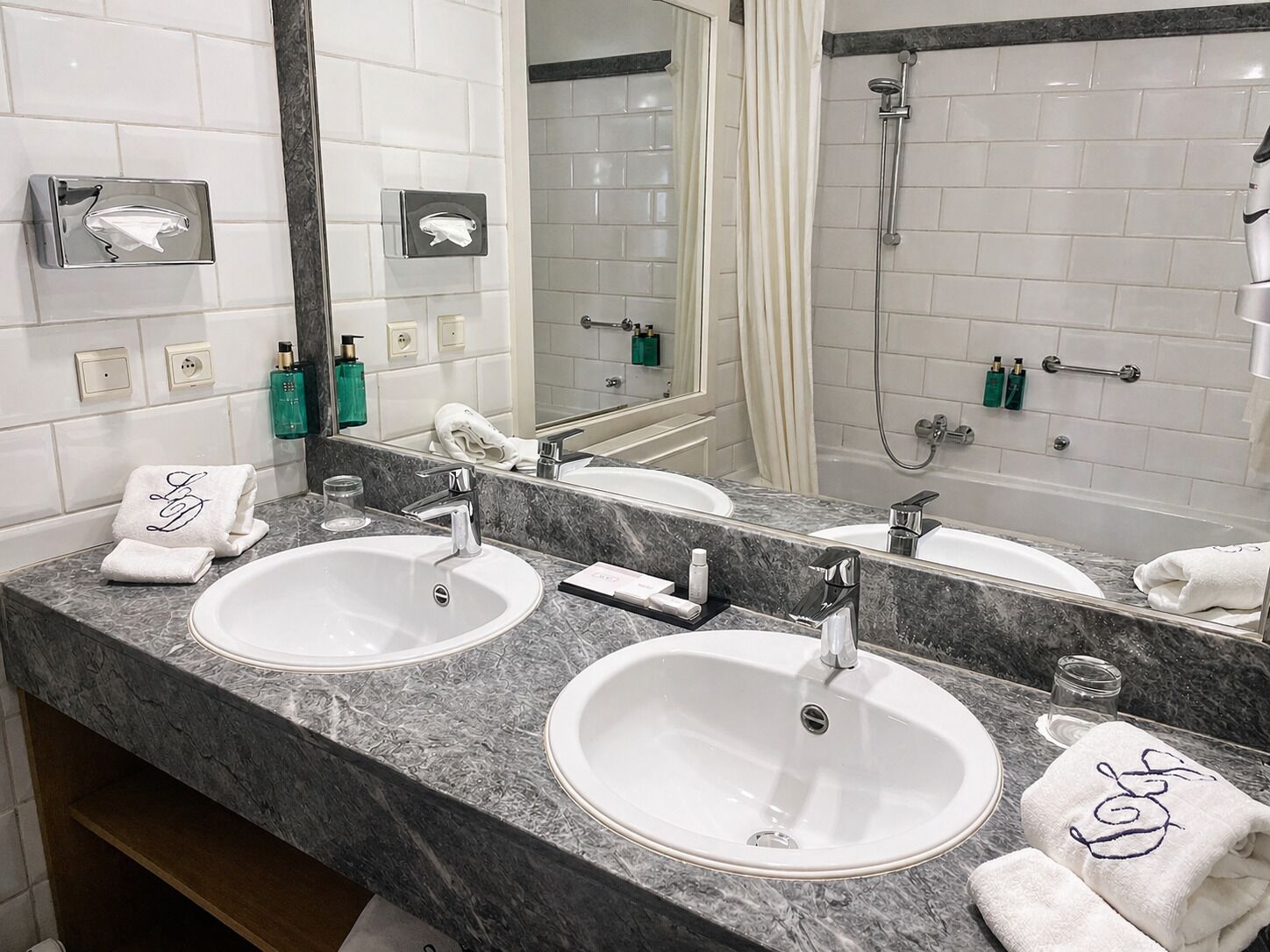 Un baño con dos lavamanos, una ducha, un perchero para toallas y un espejo.