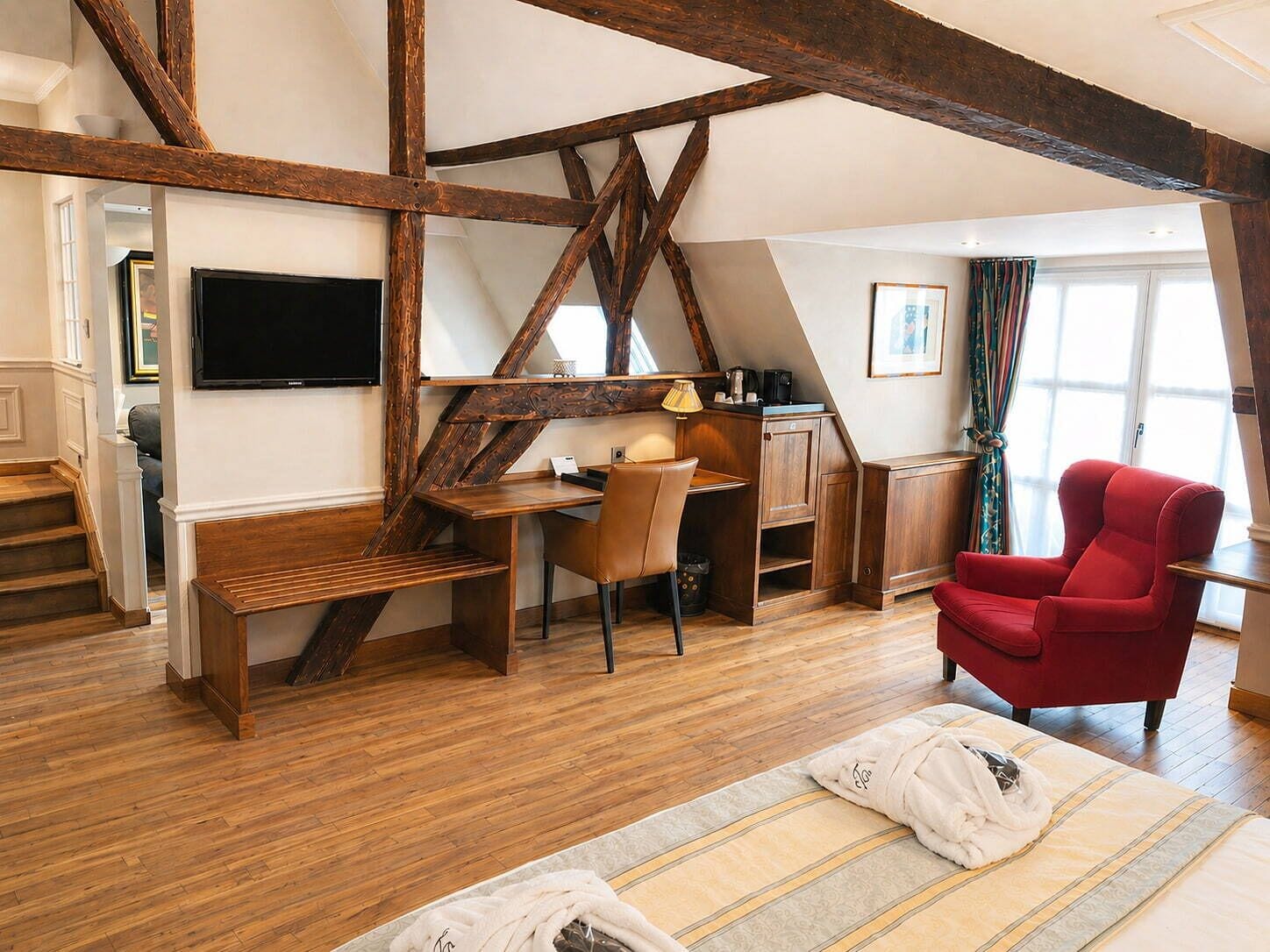 Ein Zimmer mit einem Holztisch, einem Stuhl, einem roten Sessel, einem Bett mit gestreifter Decke und einem an der Wand montierten Fernseher.