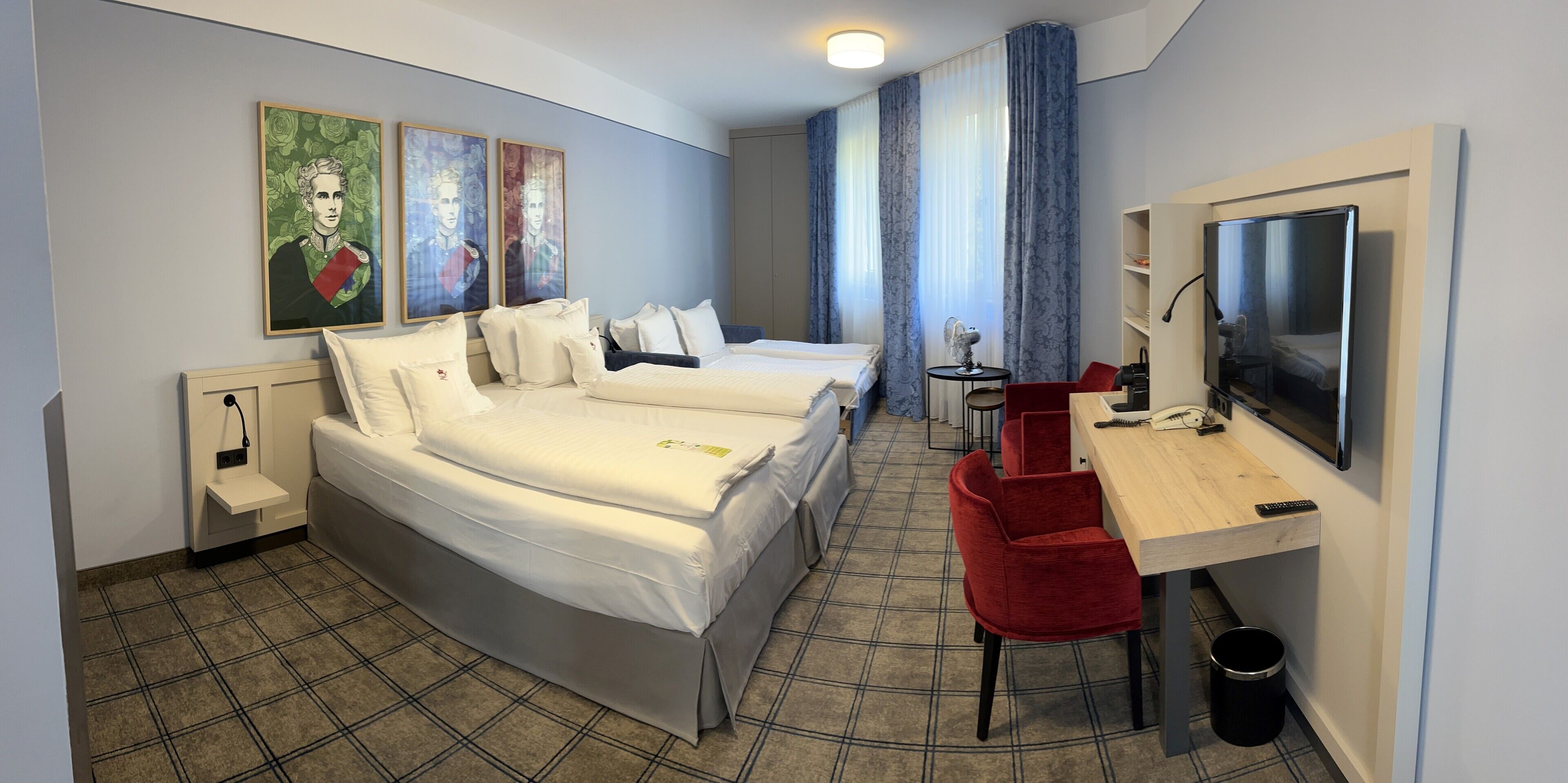 A hotel room with two beds, a desk with a TV, a chair, a small table with a lamp, and artwork on the wall.