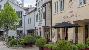 Vista de la calle del Hotel König Ludwig II, que presenta un estilo arquitectónico europeo tradicional con una entrada destacada y un jardín bien cuidado con arbustos y flores ordenados.