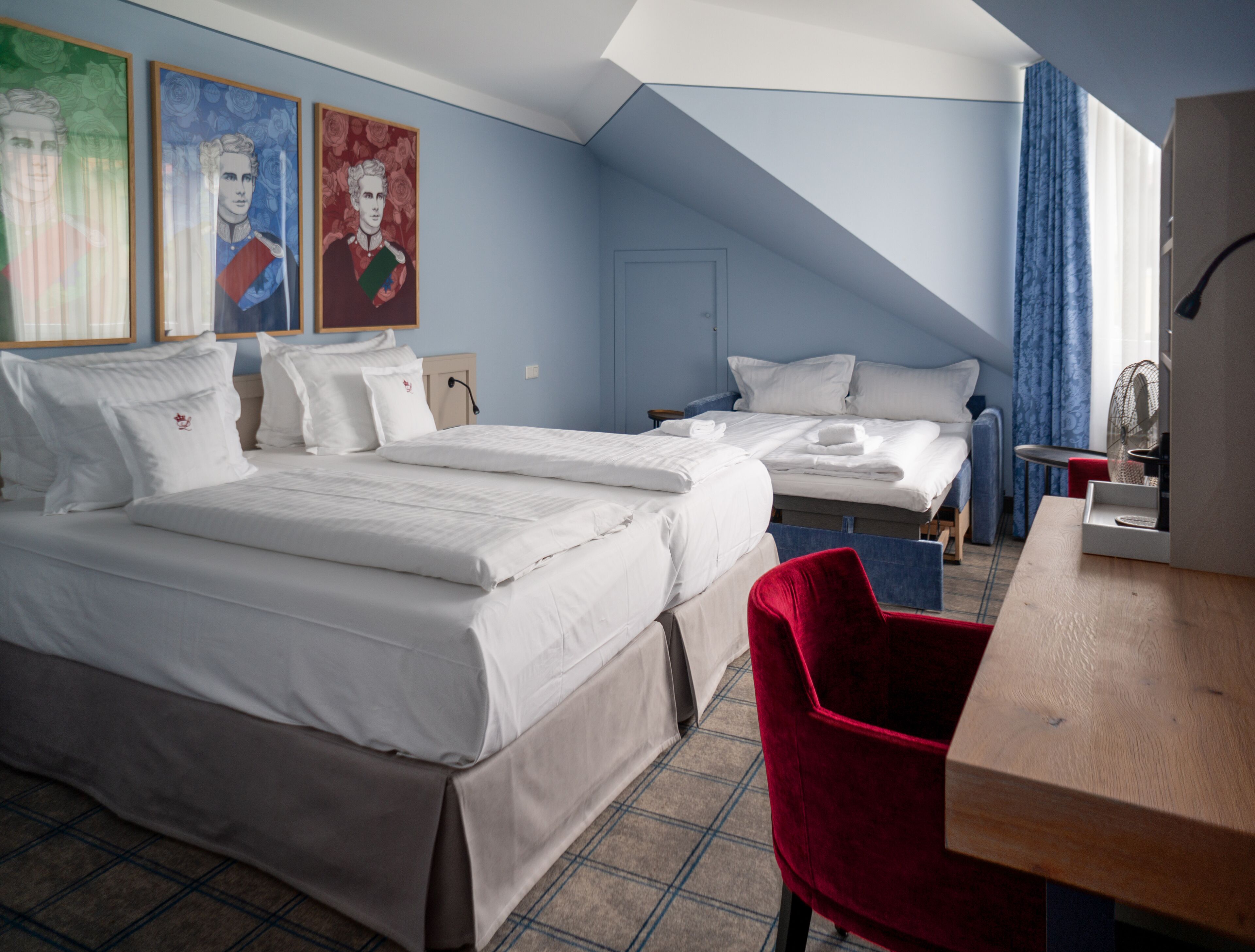 A hotel room with two beds, a red chair, a wooden desk, and a large window with curtains.