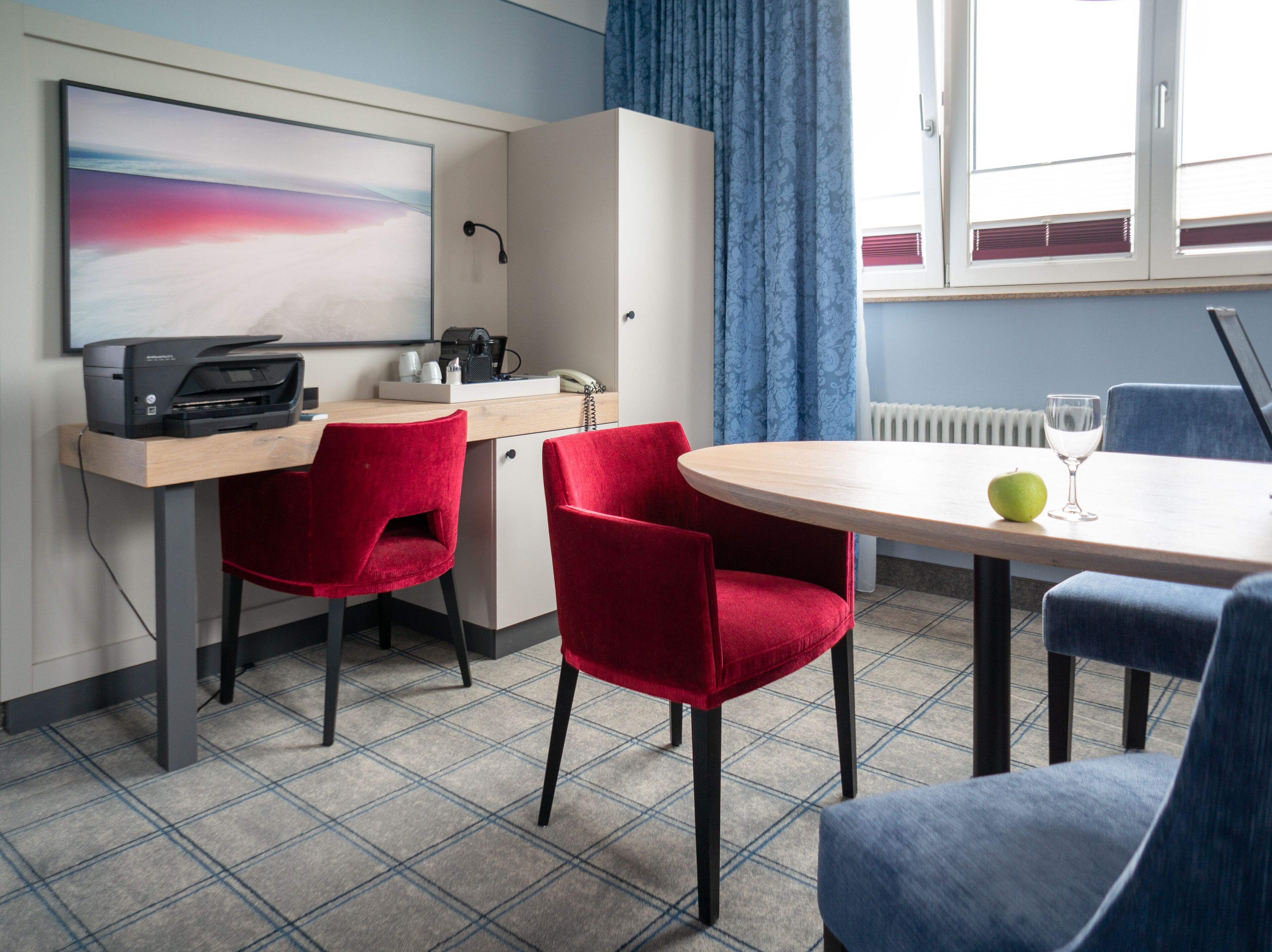 A modern hotel room with a desk, two red chairs, a round table with a glass and apple, and a large window with curtains.