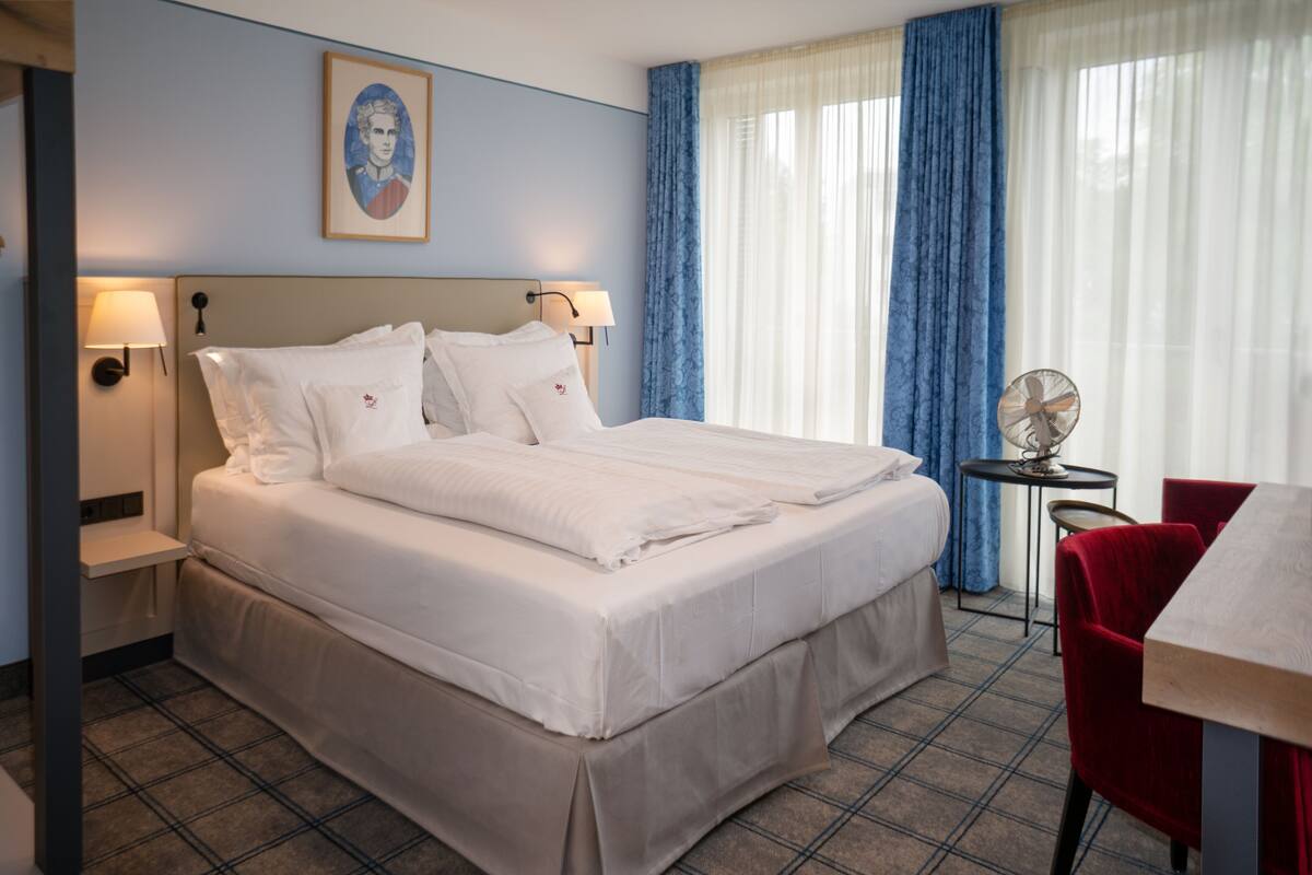 Una habitación de hotel con una cama grande, dos lámparas de noche, una mesita con un ventilador y una silla roja.
