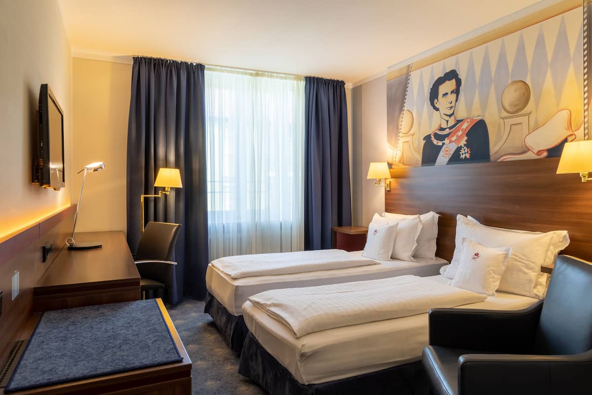 Habitación de hotel con dos camas, un escritorio, una silla, una lámpara y un cuadro en la pared.