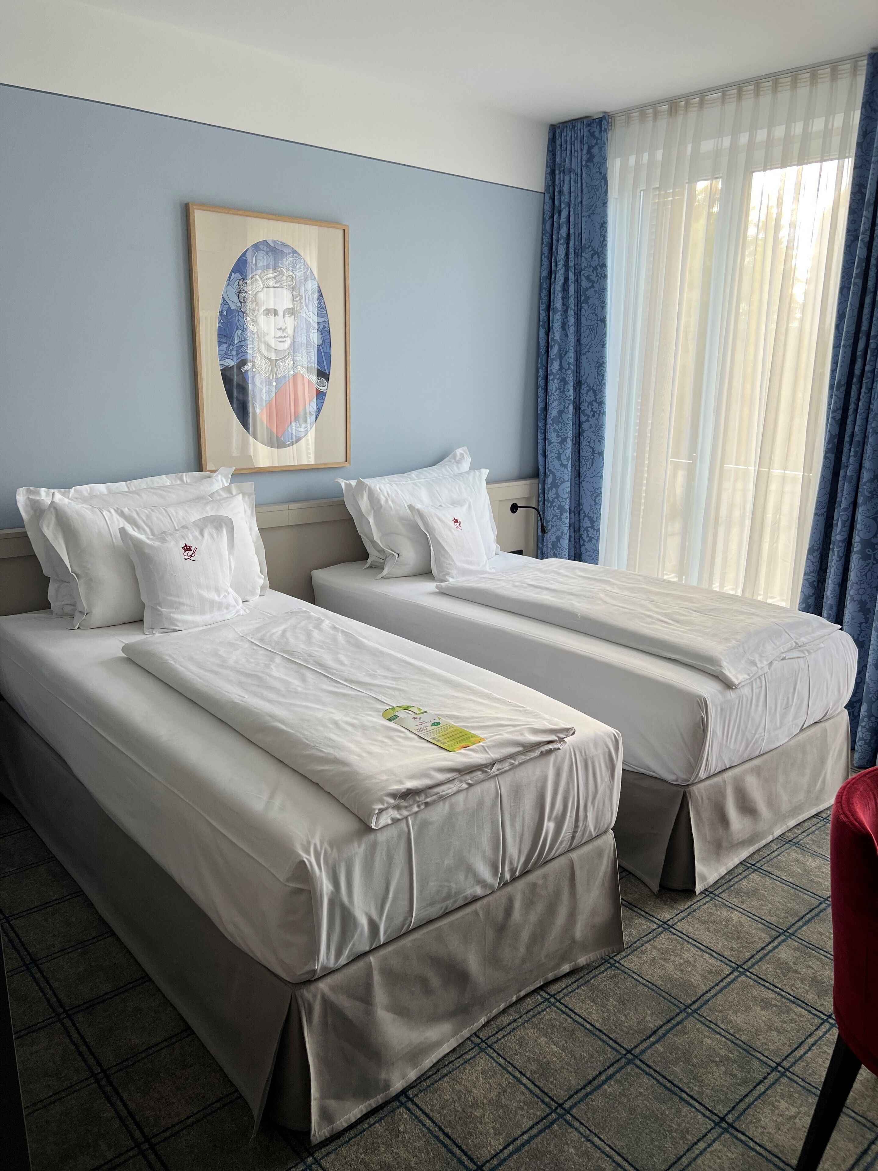 Een hotelkamer met twee bedden, een inlijst portret aan de muur en een raam met blauwe gordijnen.