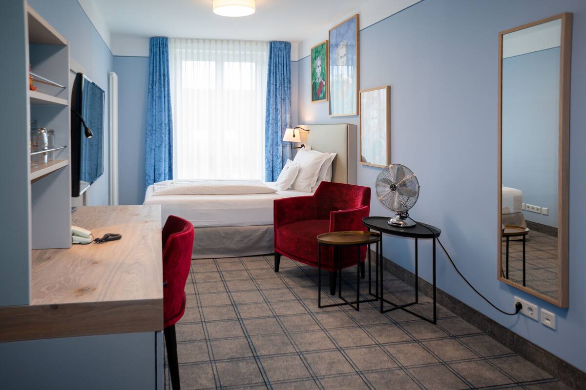 Habitación de hotel con cama, escritorio con silla, mesita con ventilador y espejo en la pared.