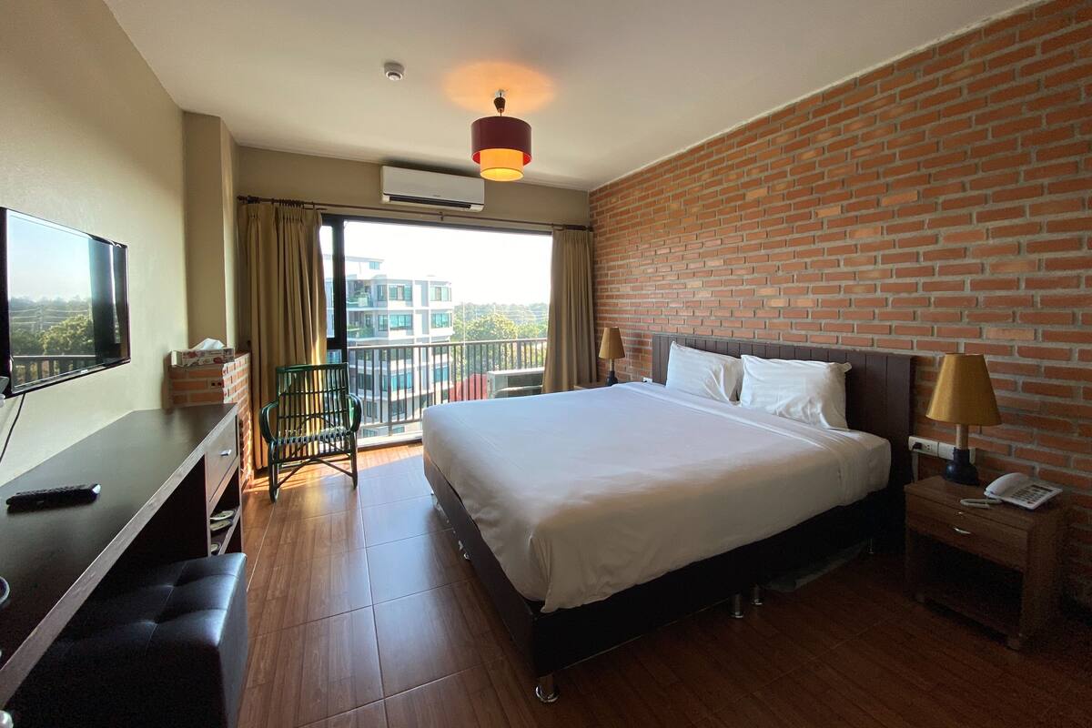 A hotel room with a large bed, a desk with a TV, a balcony with a view of buildings, and a brick wall.