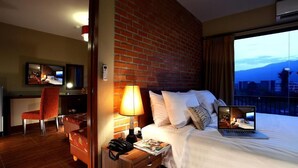 A hotel room with a brick wall, a bed with pillows, a laptop, a desk with a chair, and a nightstand with a lamp and phone. - WE Valley Hotel (Chiang Mai)