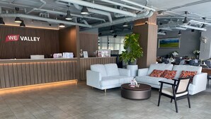 A modern reception area with a wooden reception desk, a white sofa, a round coffee table, and a large potted plant. - WE Valley Hotel (Chiang Mai)